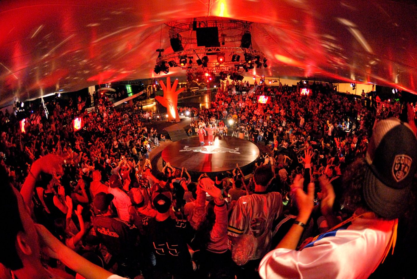 A packed arena in red light, the crowd cheering around a round battle floor.